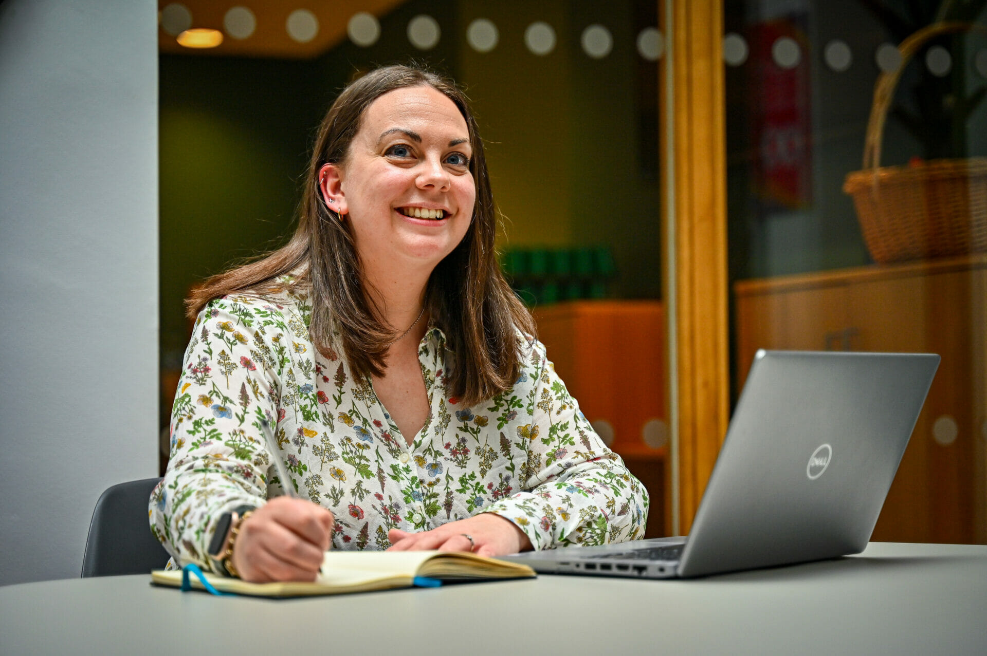 A woman smiling whilst working at a desk with a notebook and laptop in front of her.