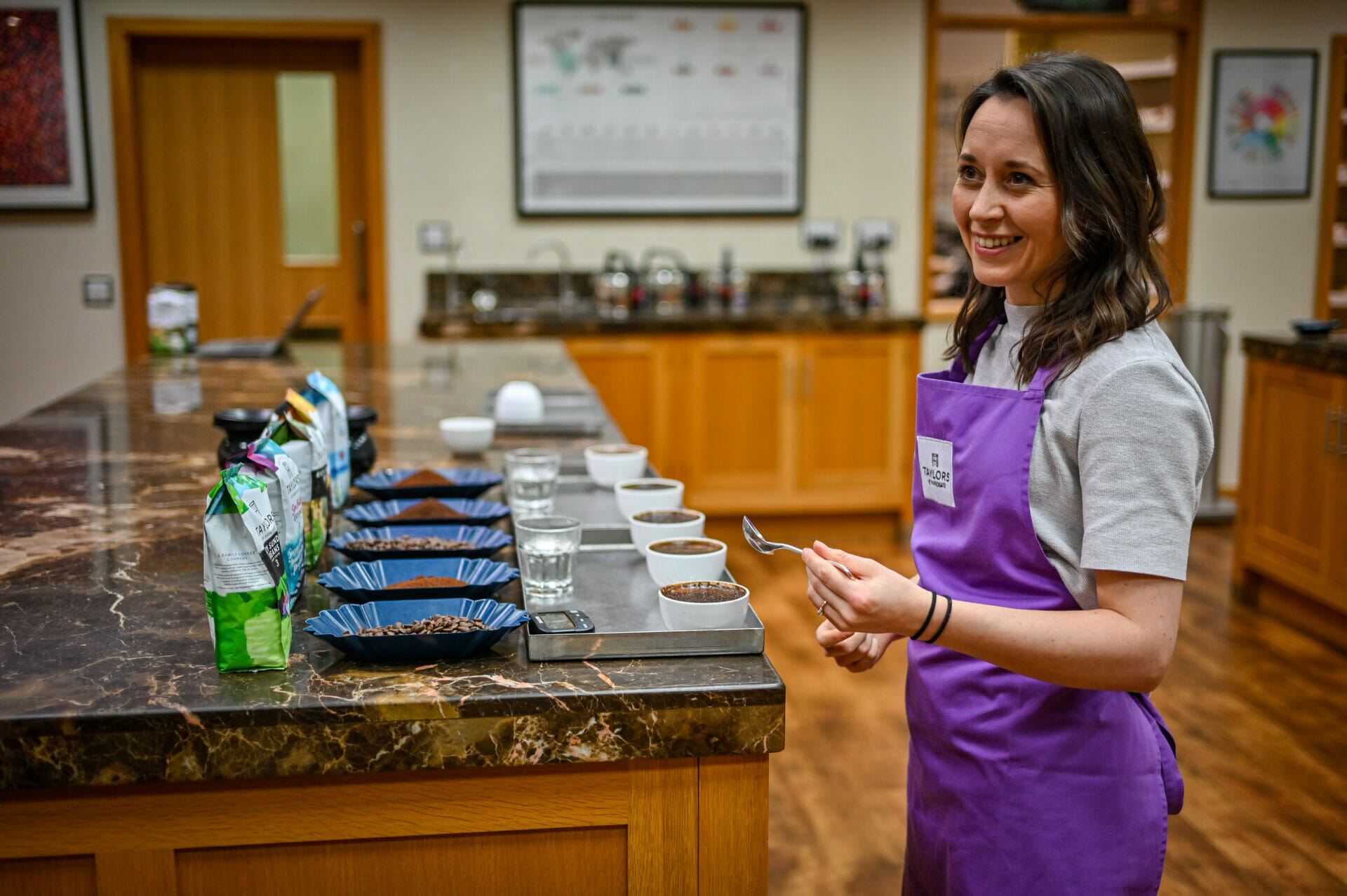 A women wearing an apron standing in front of several cups of coffee waiting to taste them.