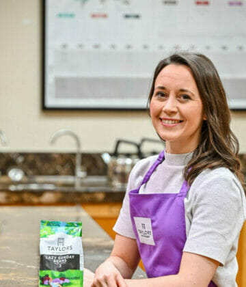 A photo of a woman learning against a table with a selection of coffee cups and trays of coffee beans laid out in front.