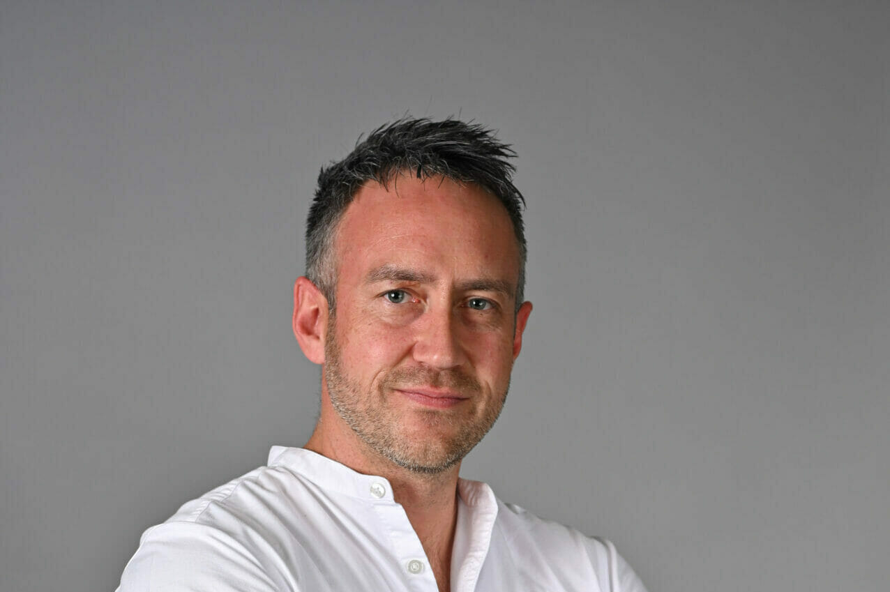 A photo of a man standing up with his arms crossed smiling at the camera.