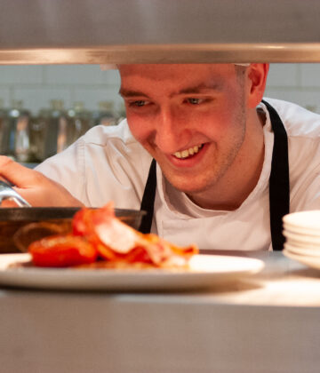 A cook serving up a dish of food in a professional kitchen to be taken out to diners.