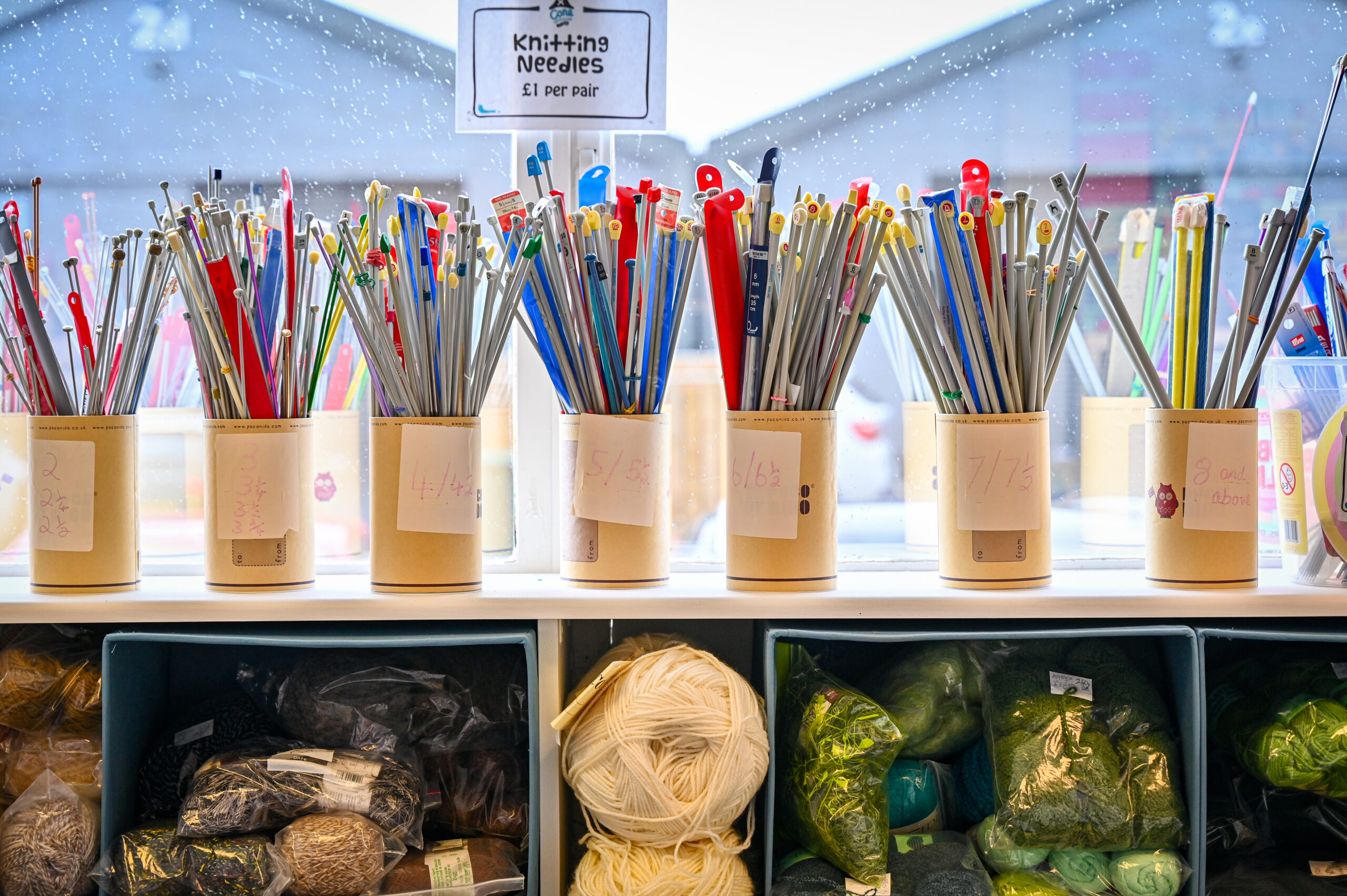 A row of pots holding knitting needles in front of a window sill, with balls of wool stored underneath.
