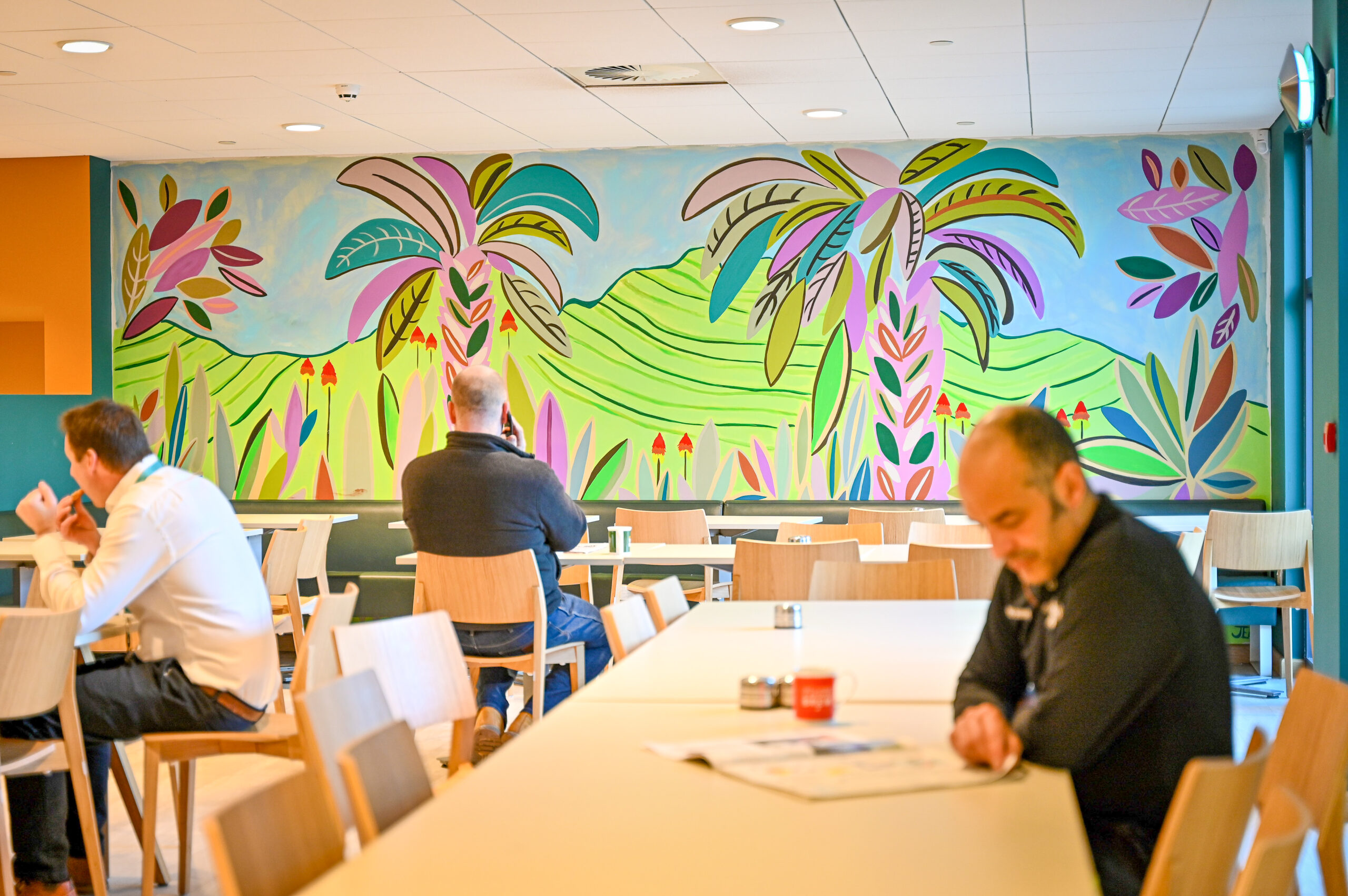 Three people seated in Taylors staff cafe, a large mural of a tropical landscape is in the background.