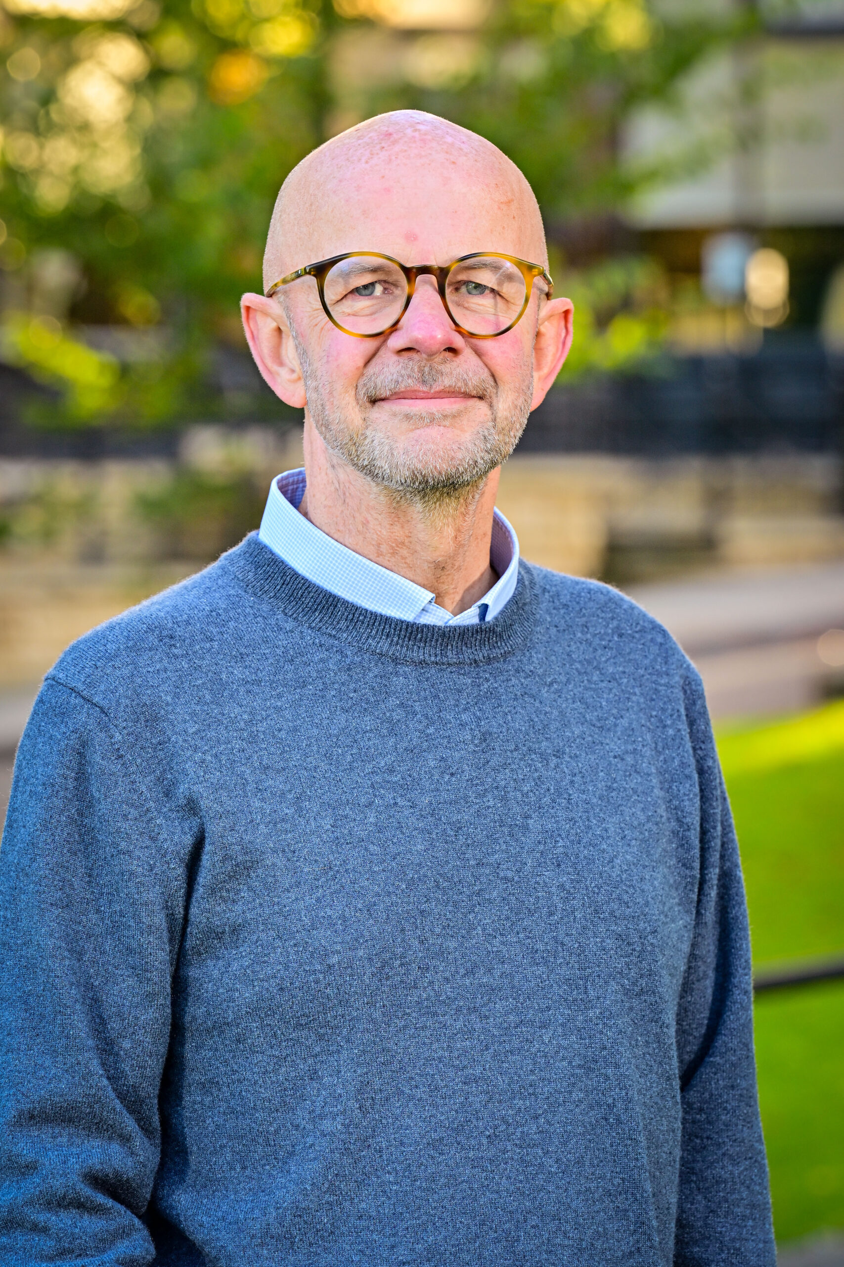 A white man wearing glasses is standing outside with the exterior of Bettys Harrogate in the background. He is smiling to the camera and wearing a pale blue shirt and a darker blue jumper.