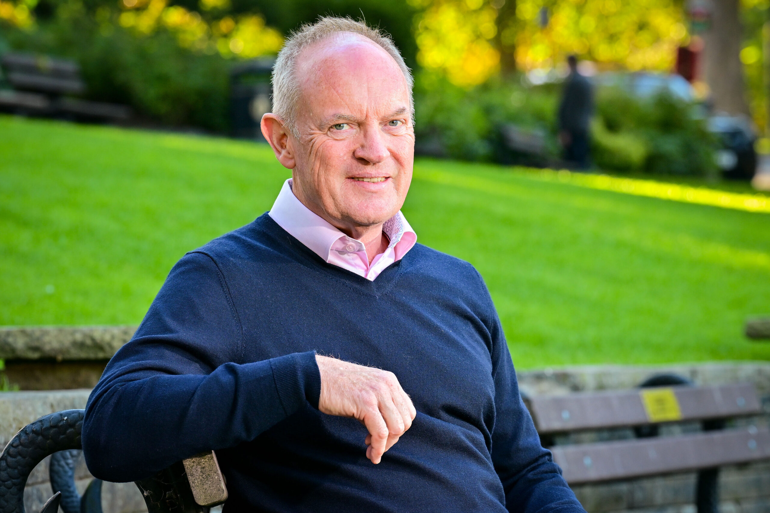 A white man wearing a pale pink shirt and a blue v neck jumper seated on a bench, a green lawn can be seen in the background.
