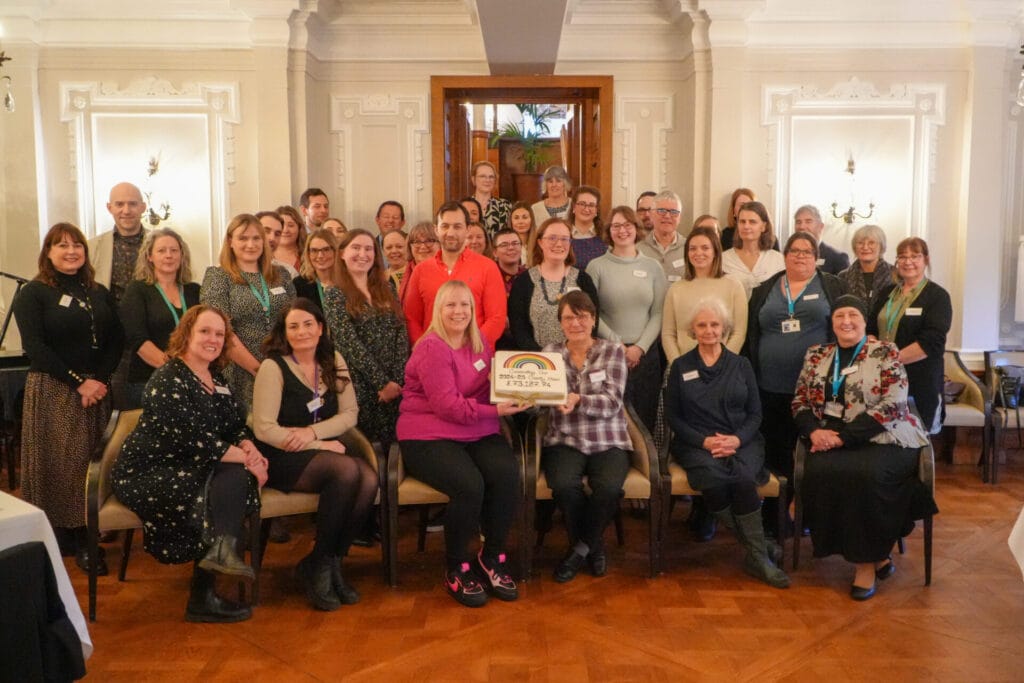 The charity representatives and charity co-ordinators are gathered in the grand Imperial Room at Bettys Harrogate. The front row of people are seated and in the middle they are holding a cake decorated with a rainbow and the total amount raised £73, 187.74 written in icing.