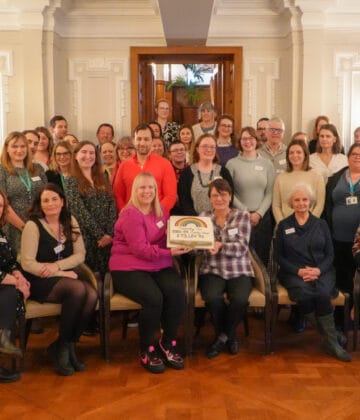 The charity representatives and charity co-ordinators are gathered in the grand Imperial Room at Bettys Harrogate. The front row of people are seated and in the middle they are holding a cake decorated with a rainbow and the total amount raised £73, 187.74 written in icing.