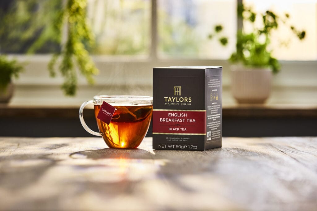 A glass tea cup holding a brewing tea with a box of Taylors of Harrogate tea next to it.