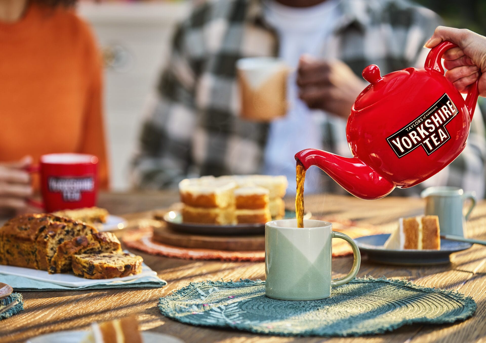 A table is laid for tea and cake. A red Yorkshire Tea tea pot is pouring tea into a pale green mug.