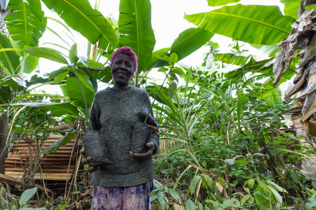 Evaryne, a TIST farmer, is holding two saplings. Behind her are large banana plants and other smaller trees.
